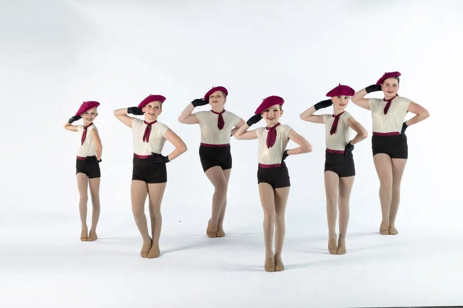 Synchronized Dance Performance with Stylish Berets A group of six young dancers performing in matching costumes, including white tops, black shorts, burgundy berets, and gloves, while striking a synchronized pose on a white background.